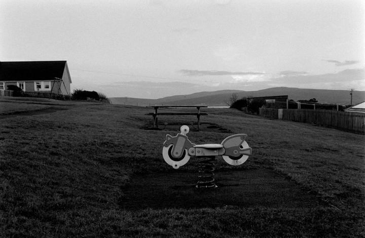 A spring rider in the shape of a motorcycle sits in a grassy field with a house and mountains in the distance.