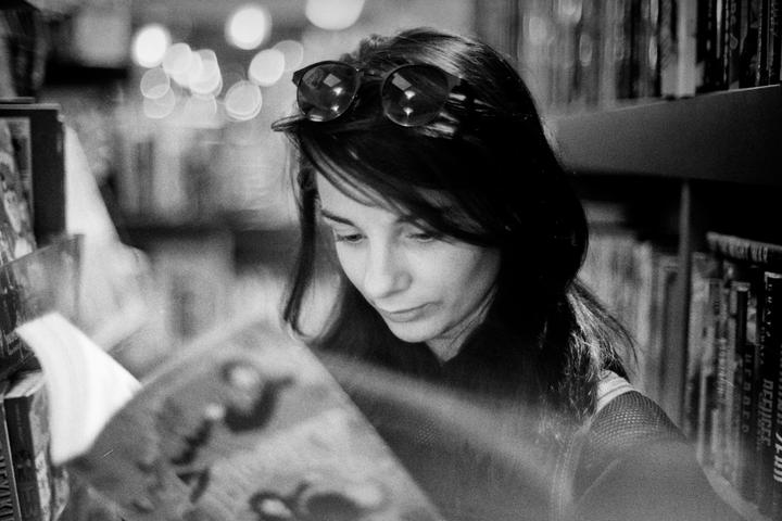 A woman with sunglasses on her head looks down at a book in a dimly lit room with shelves of books in the background.
