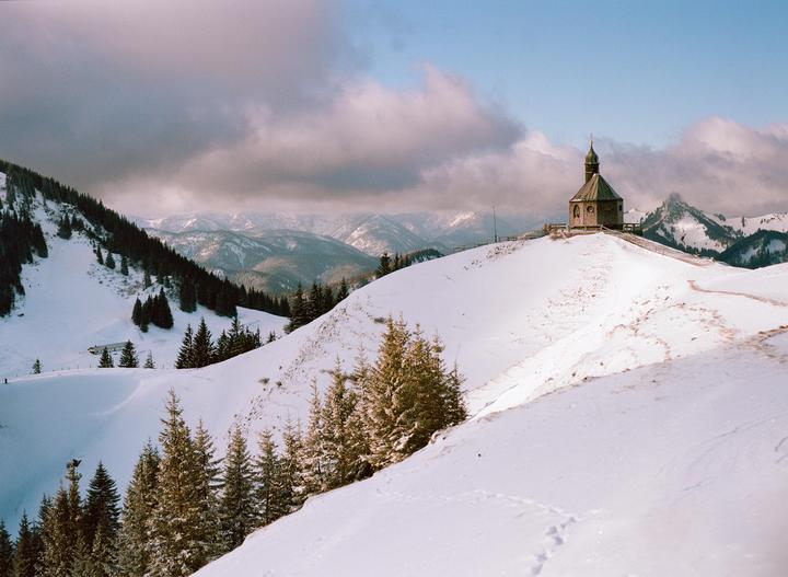 A small chapel sits atop a snow-covered hill, surrounded by pine trees and distant mountains under a cloudy sky.