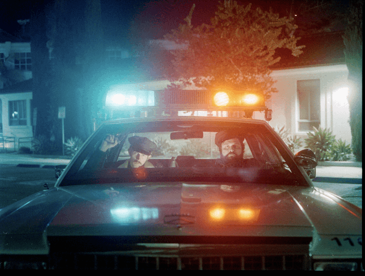 Two men in a police car with flashing lights are parked on a street at night.