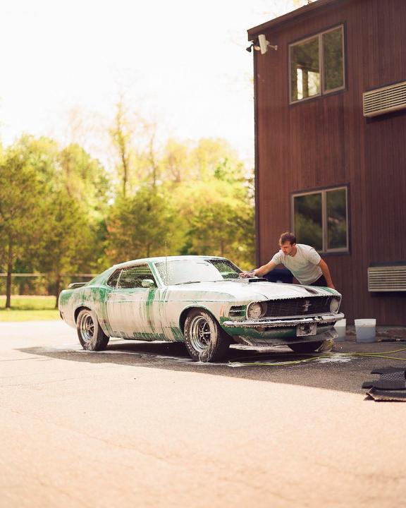 A man washes a green vintage car covered in soap suds next to a wooden building.