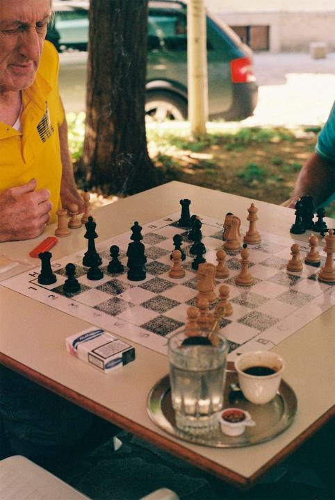 A man in a yellow shirt plays chess at a table with a glass of water and a cup of coffee.