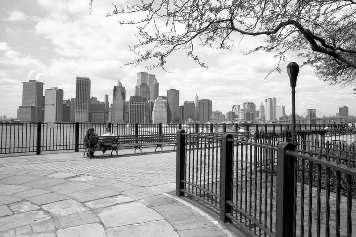 A couple sits on a bench overlooking a city skyline from a paved promenade with a metal fence and bare tree branches.