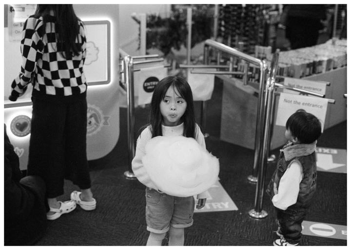 A girl with long hair holds a large cloud of cotton candy while standing in an indoor space with another child and an adult.