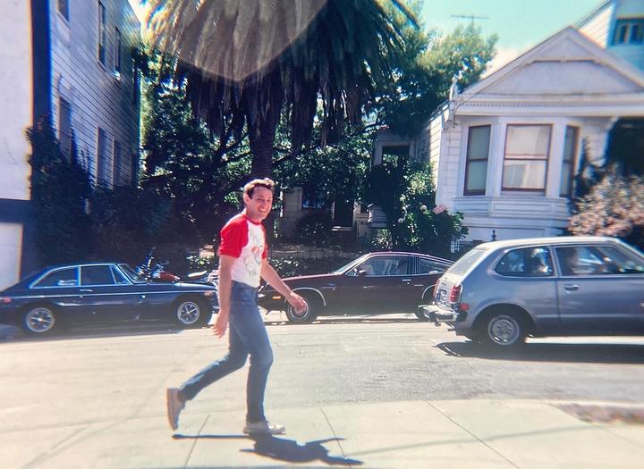 A man in a red and white shirt walks on a sidewalk next to parked cars and houses.