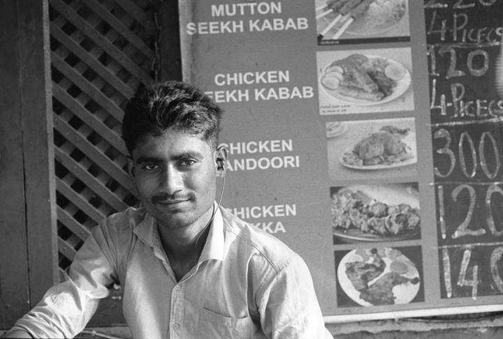 A man with an earbud smiles at the camera in front of a menu board with food items and prices.