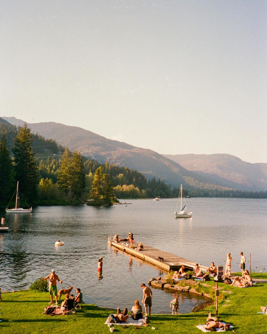 People are swimming and relaxing on a grassy shore and a wooden dock next to a lake with sailboats and mountains in the background.