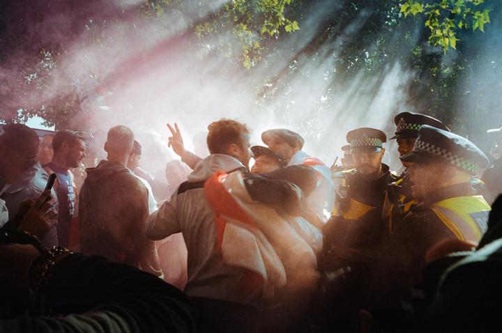 A crowd of people, including police officers, are enveloped in pink smoke with sunlight streaming through trees.