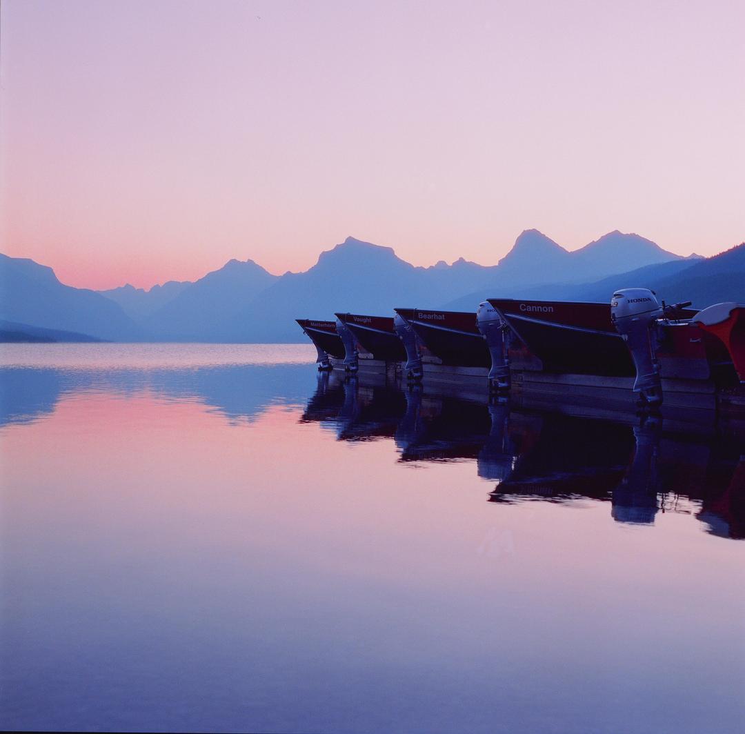 Several boats are docked on a calm lake with mountains in the background under a pink and blue sky.