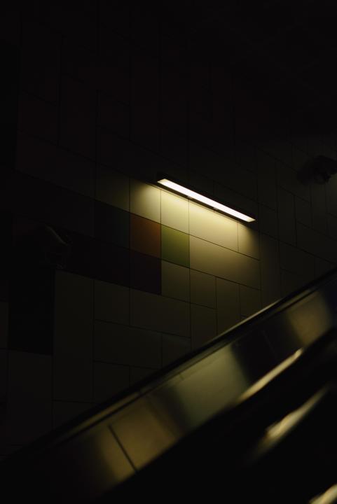 A fluorescent light illuminates a tiled wall above an escalator.