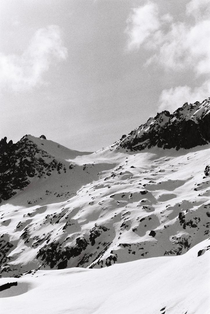 A black and white photograph captures a snow-covered mountain range under a cloudy sky.