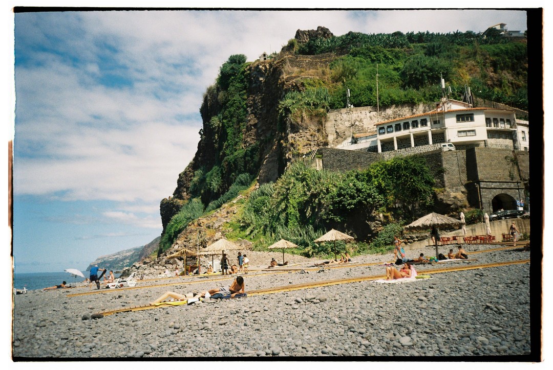 A rocky beach with people sunbathing and swimming is overlooked by a large green cliff with buildings on its side.