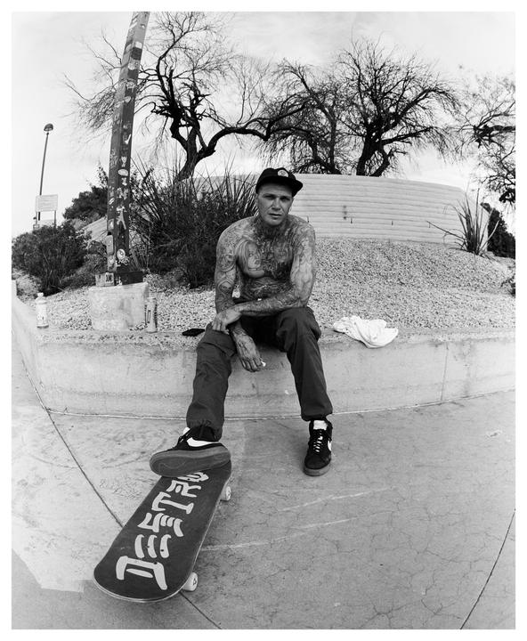 A tattooed man sits on a concrete ledge with one foot on a skateboard.