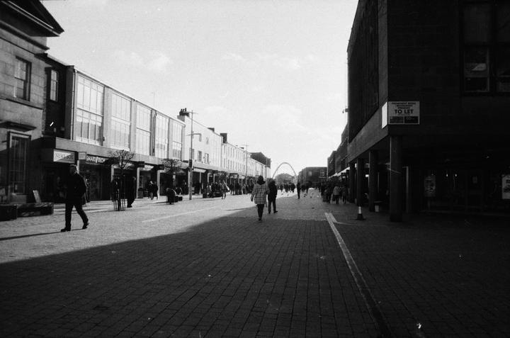 A wide street with buildings on both sides is filled with people walking, with a large archway visible in the distance.