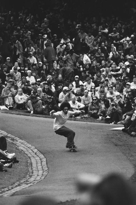 A man on a skateboard performs a trick on a paved path in front of a large crowd of spectators.