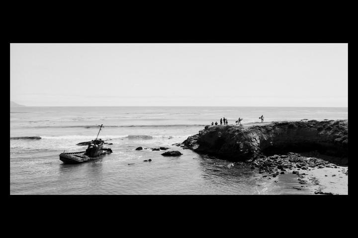 A shipwreck is visible in the water near a rocky shore where several people are standing.