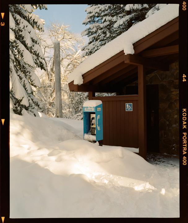 A blue payphone stands next to a brown building, surrounded by deep snow and snow-covered trees.