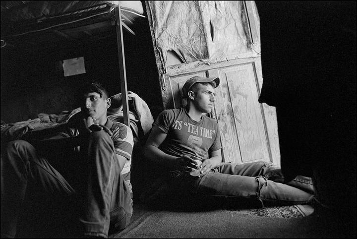 Two men sit on the floor in a dimly lit room, one wearing a cap and a t-shirt that says "TEA TIME".