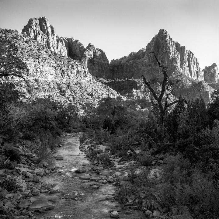 A river flows through a rocky landscape with mountains and trees under a bright sky.