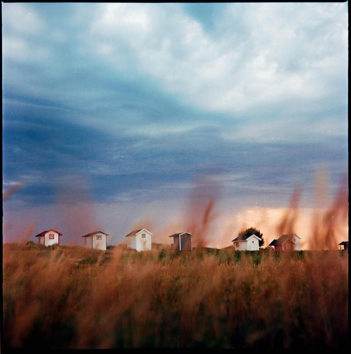 A row of small beach huts sits on a grassy hill under a stormy sunset sky.