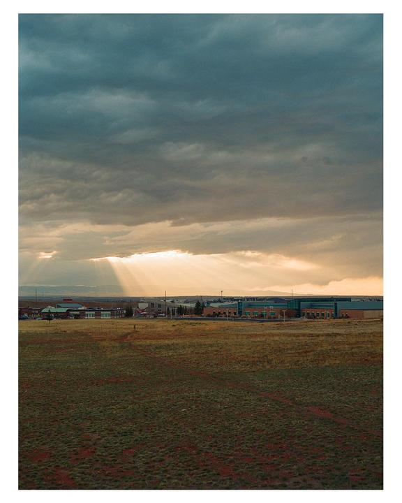 Sunlight streams through dark clouds over a field with buildings in the distance.