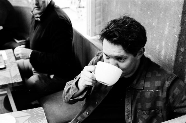 A man in a plaid shirt drinks from a white cup while another man sits in the background.