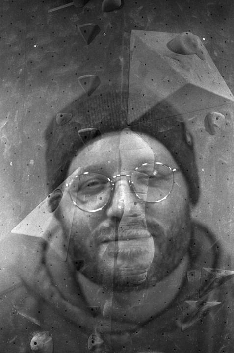 A man's face is double exposed over a climbing wall with various holds.