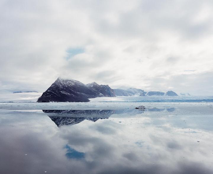 A snow-capped mountain range is reflected in a body of water under a cloudy sky.