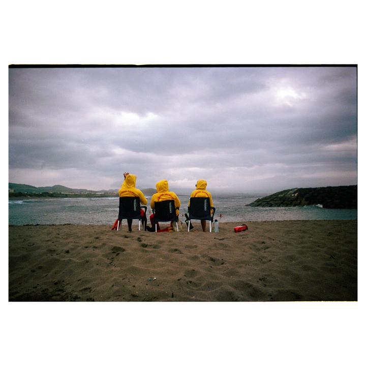Three people in yellow hooded jackets sit in chairs on a sandy beach, looking out at the ocean under a cloudy sky.