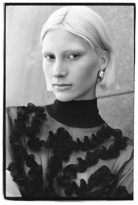 A woman with short blonde hair and a dark top with ruffles looks directly at the viewer.