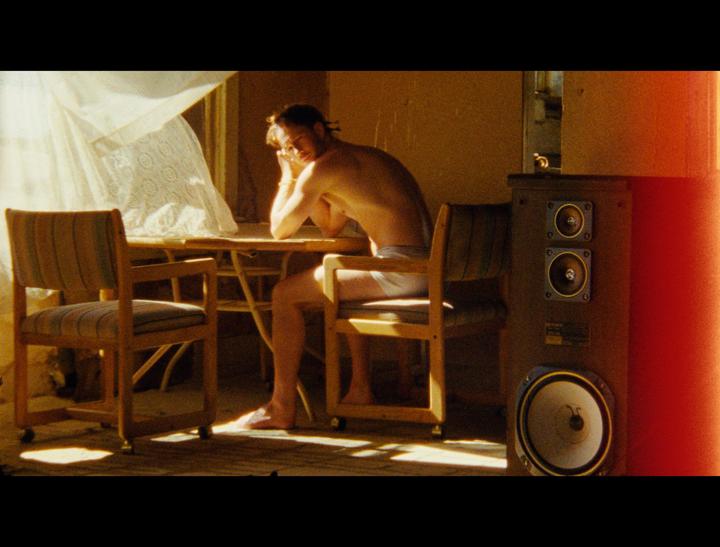 A man sits at a table next to a window with a sheer curtain and a large speaker.