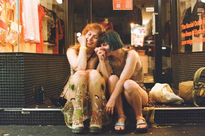 Two women sit on a tiled ledge in front of a storefront at night, holding hands and smiling.