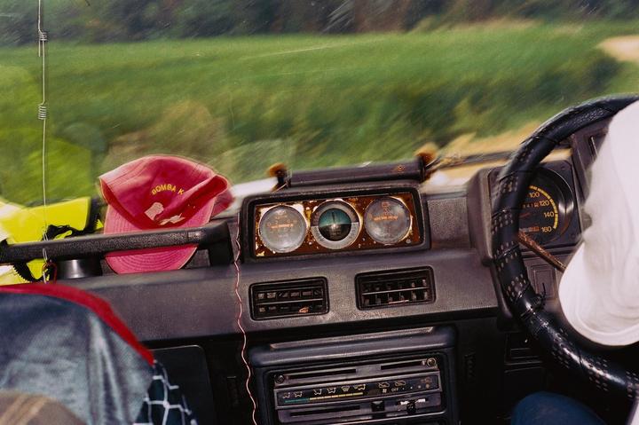 A red cap rests on the dashboard of a vehicle, with a person's arm visible near the steering wheel and a green field blurred in the background.