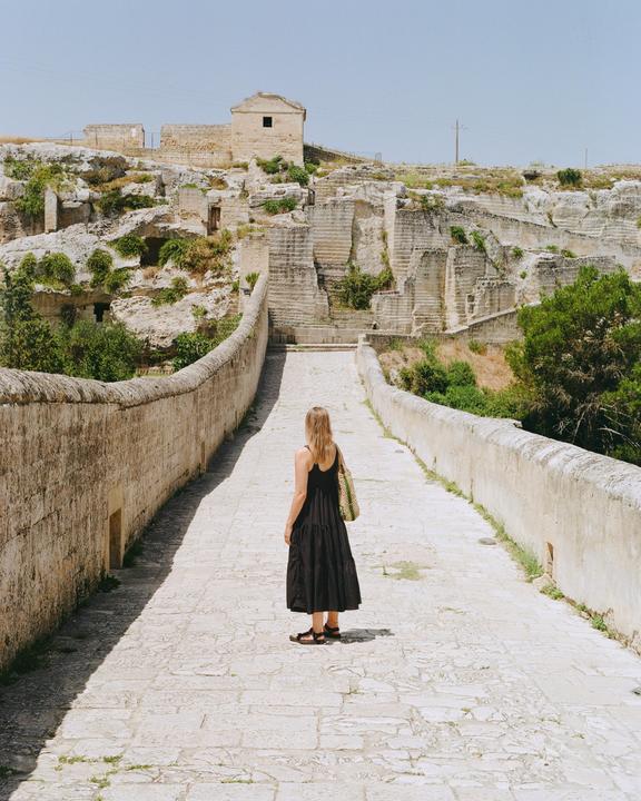 A woman stands on a stone bridge looking towards a hillside with ancient stone structures and vegetation.
