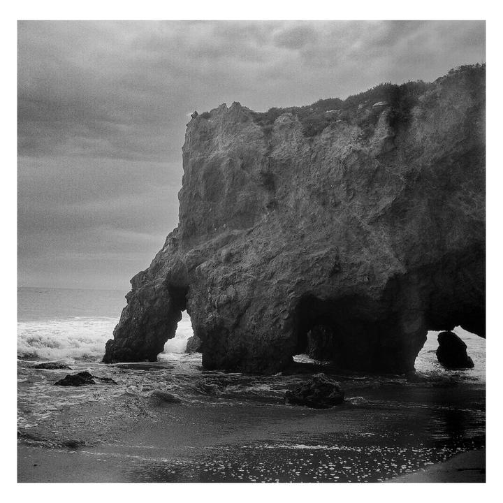 A large rock formation with natural arches stands in the ocean as waves crash on the shore under a cloudy sky.