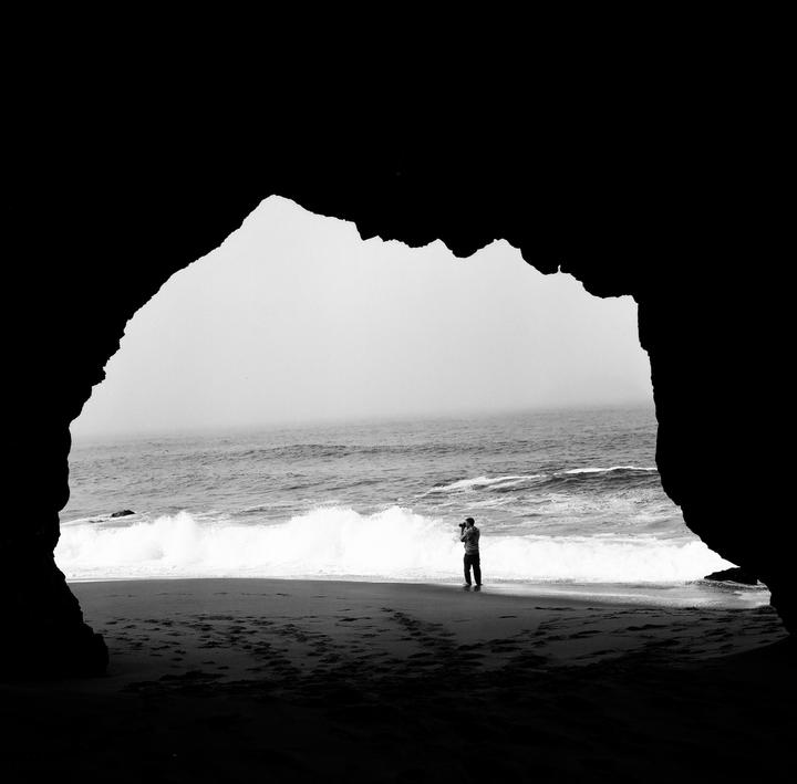 A man stands on a beach with waves crashing, framed by a dark cave opening.