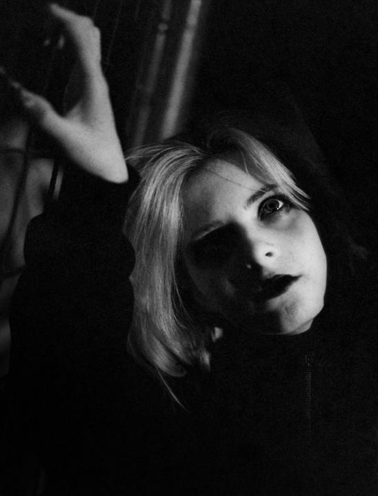 A woman with light hair and dark lips looks up with one arm raised in a dark setting.