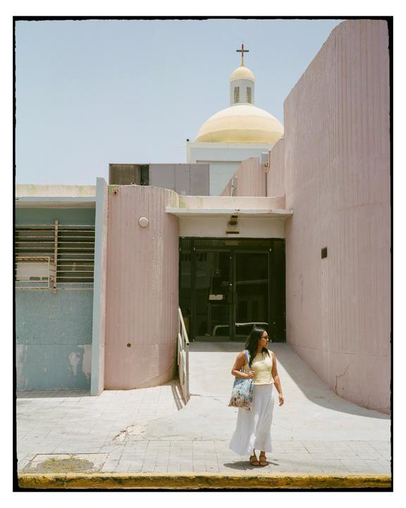 A woman stands on a sidewalk next to a pink building with a church dome and cross visible in the background.