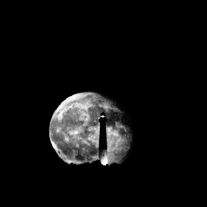 A lighthouse stands in front of a large full moon against a dark sky.