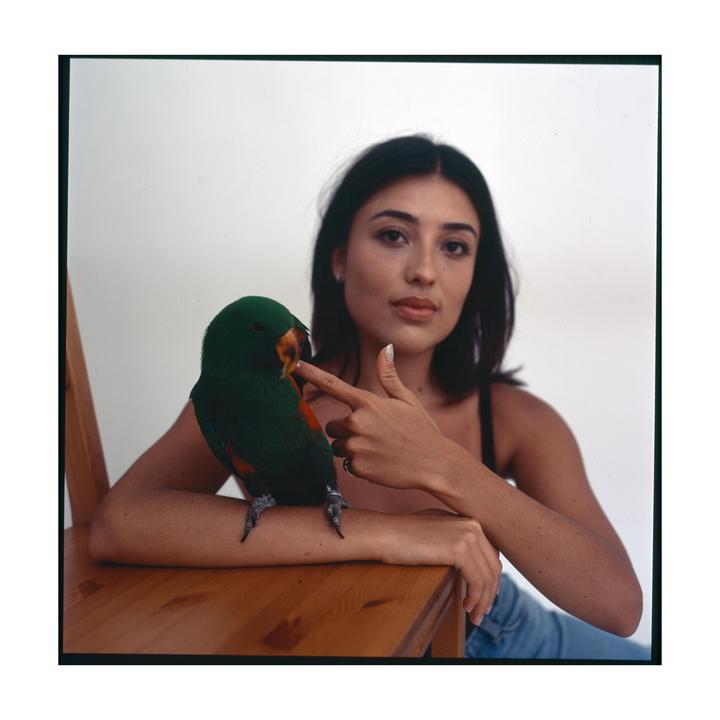 A woman sits next to a parrot perched on a wooden chair, pointing her finger at the bird's beak.