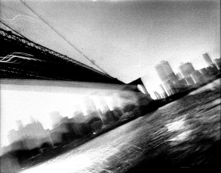 A blurry, high-contrast, black and white image shows a bridge and a cityscape reflected in water.
