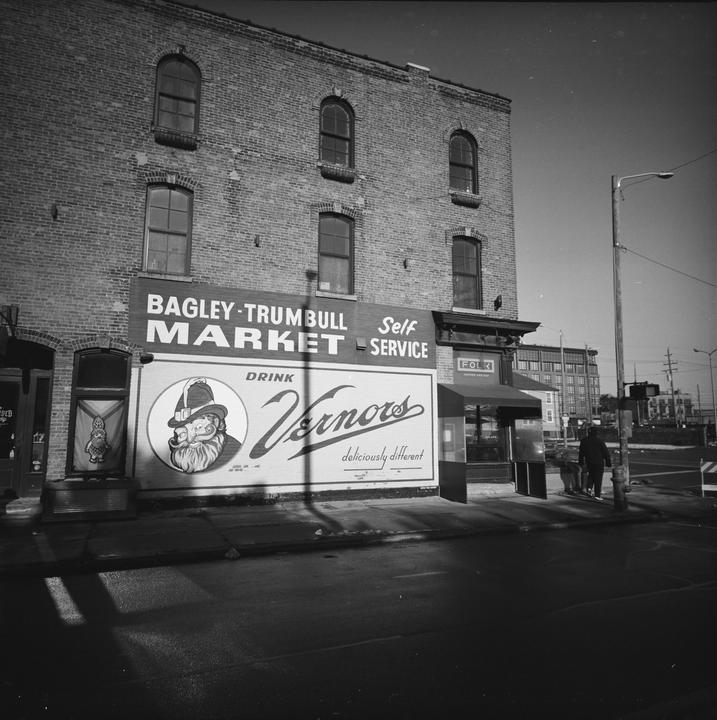 A large sign for Bagley-Trumbull Market and Vernors soda is displayed on the side of a brick building in a city.