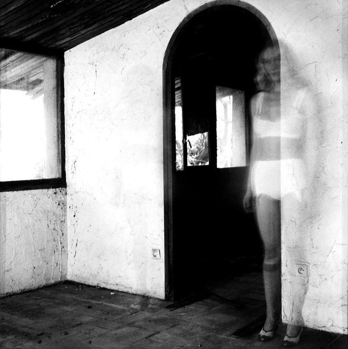 A ghostly woman stands in an arched doorway in a room with textured walls and a wooden ceiling.