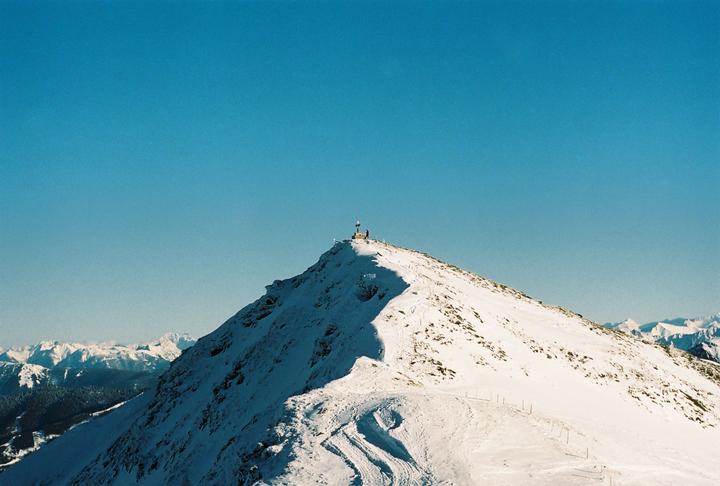 A snow-covered mountain peak with a small cross and a person at its summit is visible under a clear blue sky.