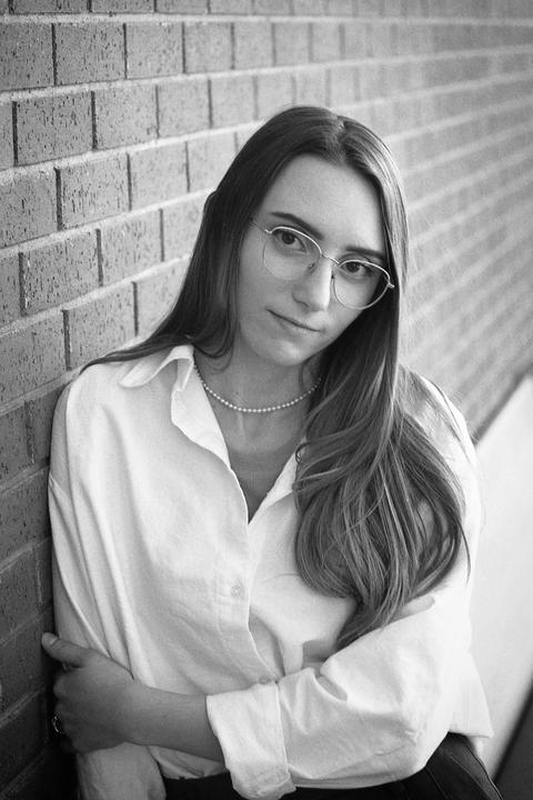 A woman wearing glasses and a pearl necklace leans against a brick wall.