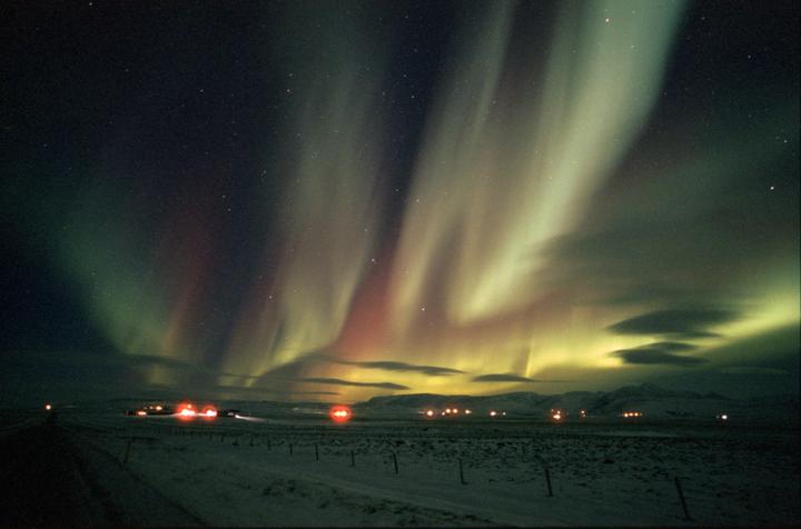 The aurora borealis illuminates a snowy landscape with distant lights and mountains under a starry night sky.