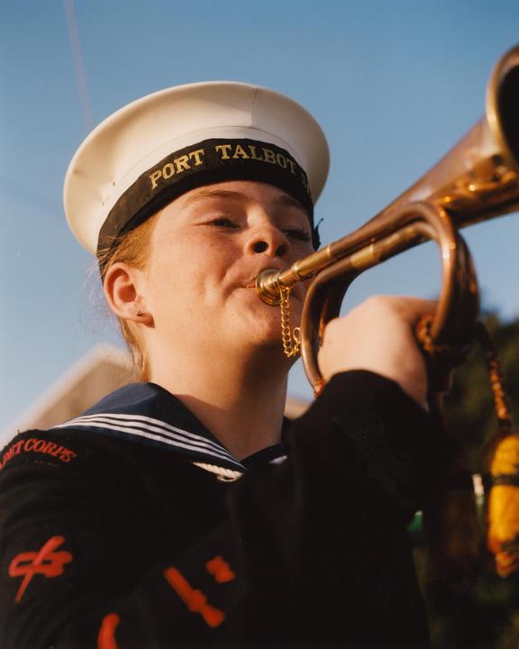 A woman in a naval uniform and hat plays a brass instrument against a clear sky.