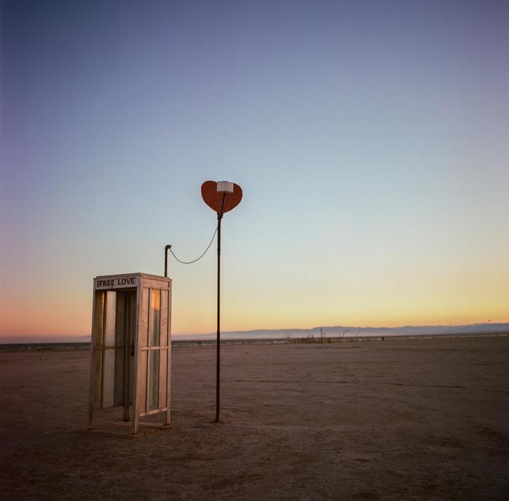 A phone booth with "FREE LOVE" written on it stands next to a pole with a heart shape at its top in a desert landscape at dusk.