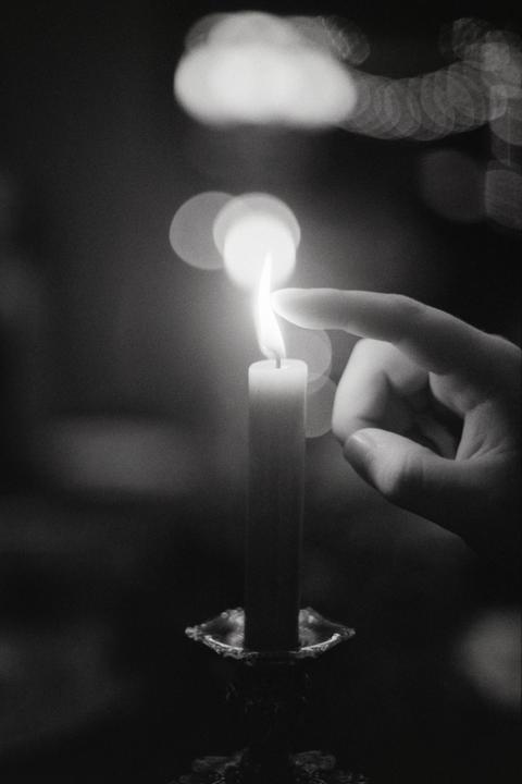 A hand reaches out to touch the flame of a lit candle in a dark setting.