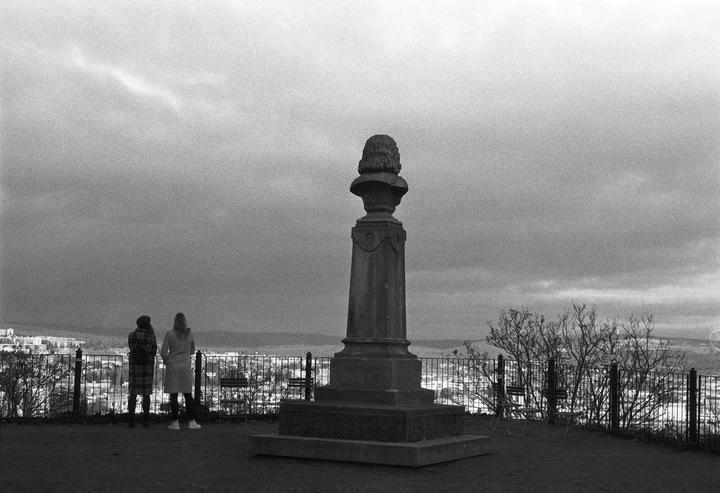Two women stand with their backs to the camera, looking out over a city from a hill with a statue.